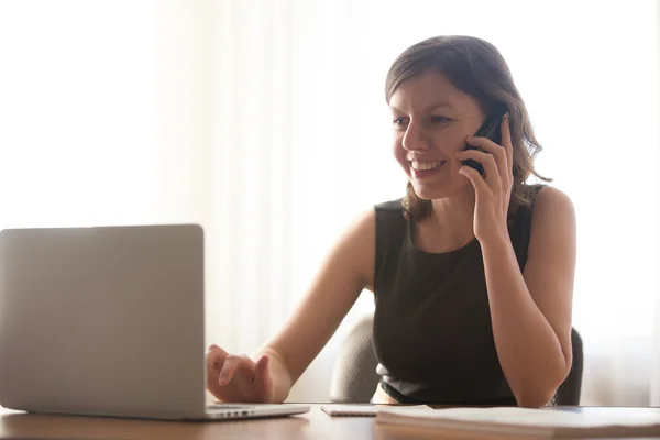 Chica joven hablando en el teléfono inteligente y escribiendo en el ordenador portátil — Foto de Stock