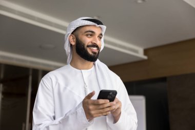 Beyaz keffiyeh 'li başarılı Arap iş adamı. Ofiste cep telefonu kullanıyor, akıllı telefonu tutuyor, başka yere bakıyor. Çevrimiçi iletişimi düşünüyor, mesaj atıyor, gülümsüyor.