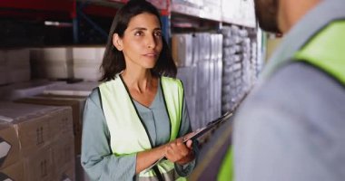 Female logistics worker in vest shaking hands with male colleague standing in warehouse aisle, surrounded by industrial shelving, boxes, and packaging materials. Teamwork, agreement, or partnership