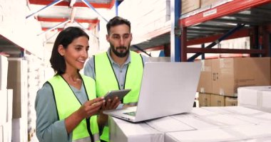 Two logistics workers collaborating in storage facility, reviewing inventory records, updating warehouse management system using tablet and laptop. Technology, stock control and shipment coordination