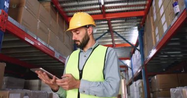 Young 30s Latino male logistics worker wearing hard hat and protective vest working on modern digital tablet, makes inventory control, updating stock information, verify shipments in logistics center