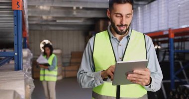 Male warehouse worker in vest using handheld barcode scanner to check inventory on shelves, while holding digital tablet, engaged in stock verification or logistics tracking in modern storage facility