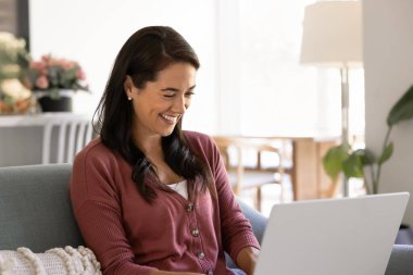 Happy young Hispanic freelancer woman working at home, typing on laptop, sitting on sofa in living room, laughing. PC user enjoying entertaining multimedia service, online application, smiling
