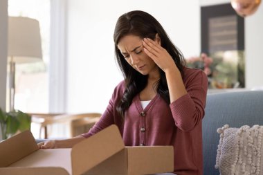 Frustrated young Latin customer woman unpacking parcel at home, touching head, feeling sick, headache, stress, frustration, looking into cardboard box, finding damage, mistake