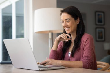 Cheerful happy young Latin remote professional woman typing on laptop at home, using Internet for business communication, browsing social media, smiling, laughing, talking in video call
