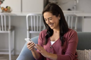 Cheerful 30s Latin woman having fun, typing on smartphone at home, enjoying leisure, communication on Internet, looking at mobile phone, watching online content, laughing