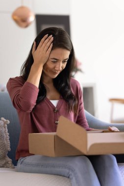 Confused disappointed young Hispanic woman opening cardboard box at home, looking at wrong damaged parcel received from delivery transportation, service, holding head. Vertical shot