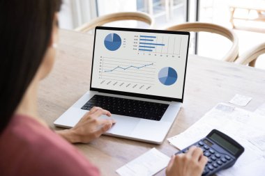 Over shoulder view of laptop screen with analytic information on table of freelance business woman analyzing economic growth, product sales benchmarks, using calculator