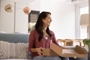 Happy young Hispanic consumer woman holding open logistic cardboard box at home, unpacking parcel received from courier service, sitting on sofa with package, looking away, smiling
