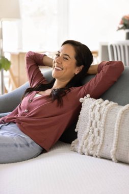 Happy cheerful young Latin woman relaxing on soft couch at home, resting on sofa, looking away with toothy smile, laughing, enjoying leisure, relaxation, comfort, coziness
