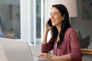 Happy young Hispanic woman speaking on cellphone at home workplace with laptop, enjoying mobile phone call, conversation, wireless communication, smiling, laughing