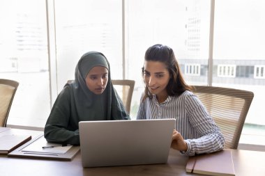 Internship and cooperation. Confident young Latin woman team leader boss expert mentoring consulting Arab female coworker in traditional Muslim wear help intern show work in corporate app on notebook