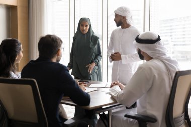 Collaboration on project. Confident multiethnic team corporate workers executives discuss work problems at meeting table listen to young Muslim woman in abaya offer business solution express idea plan
