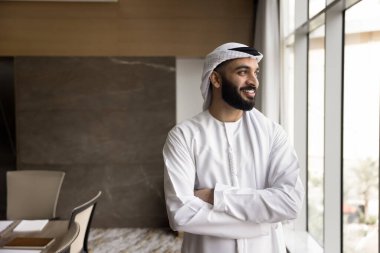 Looking forward with confidence. Young smiling Arabian man leader ceo wearing Muslim kandora stand by office window visualize happy future of own business create financial growth strategy. Copy space