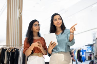 Multiethnic female retail store assistants working on sales floor together, using digital tablet. Fashion shop staff discussing merchandise for new collection. Manager instructing associate