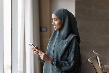 Mobile phone app. Smiling pensive young Muslim female of Arabic ethnicity wearing abaya stand by window at business office hold cell think on message text to send client in chat relax surfing websites