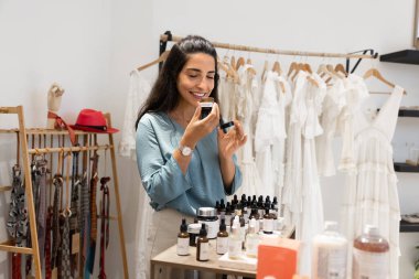 Positive relaxed young Hispanic customer woman inhaling aroma of organic cream, essential oil, keeping open jar at face and nose with closed eyes, choosing natural cosmetic product in eco boutique