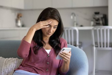 Puzzled Latin smartphone user woman touching head, reading message on mobile phone, looking at screen in shock, surprise, getting problems, financial loss from online bank account