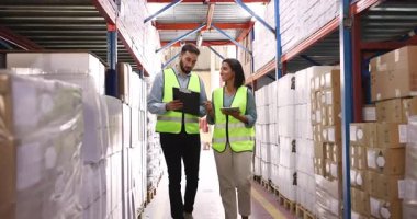 Colleagues in protective vests working together in warehouse, reviewing boxes and packages on shelves, checking inventory or logistics data on clipboard and tablet, collaborate on storage organization