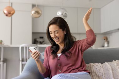 Cheerful excited Hispanic smartphone user woman celebrating online win, triumph, victory, looking at mobile phone, making surprised winner hand, laughing, enjoying success
