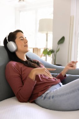 Happy inspired peaceful young Latin woman in wireless headphones resting on home sofa, listening to music, singing along songs with closed eyes, using multimedia service. Vertical shot