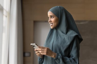 Dreamy smiling young Arabic girl student entrepreneur employee stand in office hold phone look away distracted from screen networking online read message in chat create comment text for post in blog