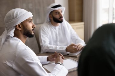 Focused on business. Concentrated young Arab businessmen wear white Muslim dress sit by conference table listen to partners at negotiations plan investment think on buying selling patent. Copy space