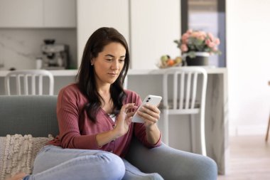 Positive young Hispanic woman using Internet technology, e-commerce platform for online shopping on cell phone, relaxing on sofa at home, typing message on smartphone