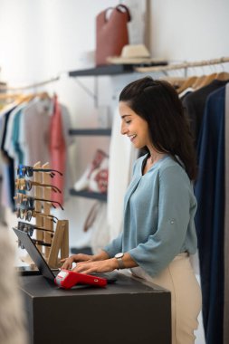 Happy Hispanic retail store manager checking sales records, stock, order processing on computer at checkout counter in fashion shop area, typing on laptop with POS terminal. Vertical shot