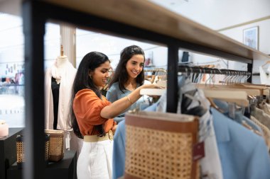 Happy young diverse retail store colleagues arranging merchandise, making inventory together. Fashion shop customer and assistant viewing clothes on rack for trying on and buying. Candid shot