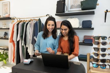 Two positive multiethnic retail store managers using computer at cash register desk, checking sales records, stock, transactions, working together, giving each other help, support