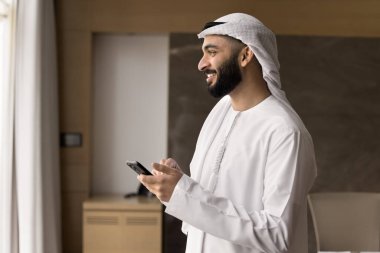 Wireless connection. Smiling young Arabic man in traditional Islamic white robe student employee relax in office with phone text message in chat play web game communicate online using wifi. Copy space