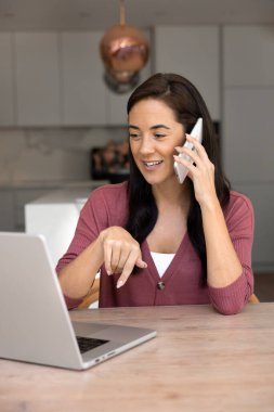 Positive busy focused Latin freelance business woman working from home, talking on cell, using company application, database on laptop for giving consultation on cellphone. Vertical shot