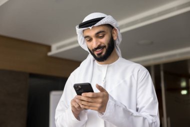 Using phone. Smiling young middle eastern businessman in Muslim white thobe keffiyeh hold smartphone read message text with good business news enjoy successful crypto currency trading in mobile app