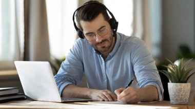 Attentive remote student. Focused millennial male in digital headphones with mic attend virtual lecture via elearning app on notebook write take notes after teacher tutor study academic subject online