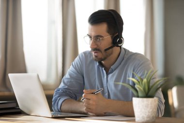 Online business meeting. Confident young man remote worker executive freelancer wear headset with microphone sit at home office desk communicate by video conference app on notebook ready to take notes