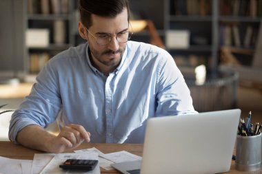 Paying bills online. Concentrated millennial businessman accountant bookkeeper use bank app on notebook to make payments calculate taxes check receipts invoices before pay via virtual banking service