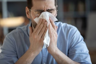 Feeling sick. Close up portrait young adult man in glasses fallen ill with cold flu influenza virus suffer from spring pollen allergy attack sneeze cough cover nose mouth with hygienic paper napkin