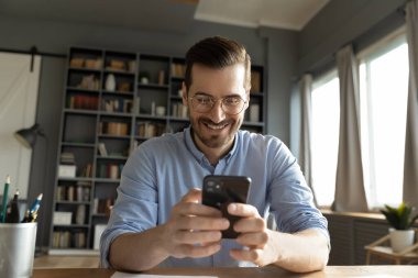 Mobile communication with smile. Cheerful glad young adult male remote worker student freelancer checking smartphone app at home office exchange friendly text messages in chat satisfied with good news