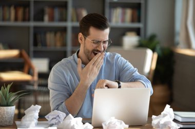 Late night deadline pressure. Tired overworked young businessman freelancer yawn check time on wristwatch struggle with sleep at home office desk by laptop with crumpled papers finish urgent project