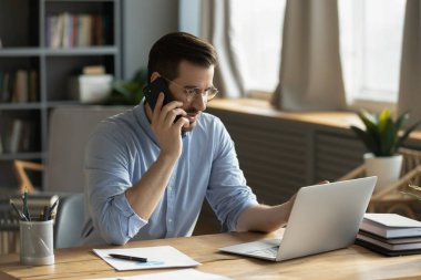 Remote multitasking. Busy millennial male business specialist project coordinator freelancer consult client by phone from home office discuss task share idea with coworker looking at notebook screen
