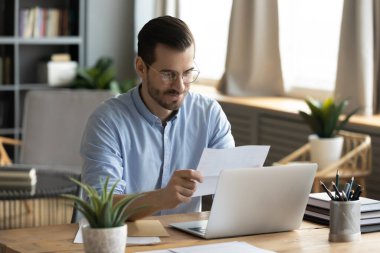 Good news from correspondence. Confident young businessman sit at workplace before notebook read paper letter received by mail with light smile feel satisfied with information from official document