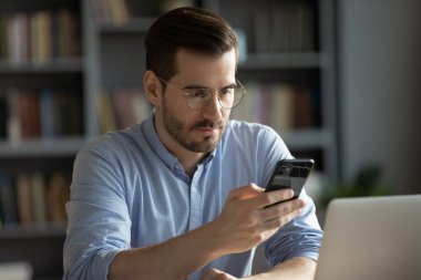 Workplace connection and communication. Focused young male business professional sit at desk by notebook manage work process using smartphone send text messages in chat check emails share information