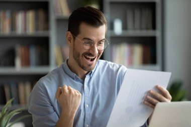 Triumph in graphs and diagrams. Overjoyed excited young businessman brand manager data analyst raise fist make yes gesture reviewing financial reports celebrate success sales growth profit achievement