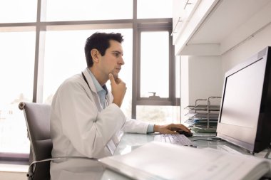 30 'lu yaşlardaki genç erkek doktor masasında oturup bilgisayarla çalışıyor, odaklanmış ve düşünceli bir ifadeyle ekrana bakıyor, tıbbi kayıtları inceliyor, test sonuçlarını analiz ediyor, ya da hasta verilerini sisteme giriyor.