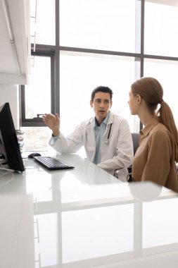 Genç pratisyen hekim veya cerrah çalışma masasında oturup kadın hastalarla konuşuyor, röntgen veya MRI görüntüleri gösteriyor, tanı ve tedavi planı sağlıyor, ilaç veya terapi taslağı hazırlıyor