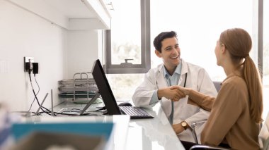 Beyaz önlüklü gülümseyen erkek doktor, profesyonel sağlık danışmanlığı randevusu sırasında modern sağlık ofisindeki bilgisayarla masada oturan mutlu kadın hastayla el sıkışıyor. Hizmet, güven