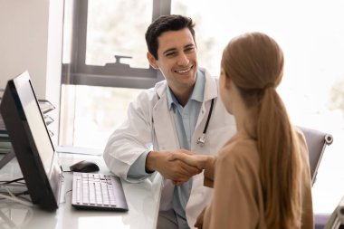 Beyaz önlüklü, steteskoplu bir terapist modern ofiste bilgisayarlı bir kadın hastayla el sıkışıyor. Tıbbi danışma, doktor-hasta ilişkisi, sağlık hizmetleri, klinik hizmetler