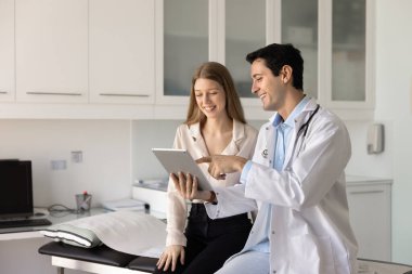 Beyaz önlüklü arkadaş canlısı erkek doktor tarama görüntülemesi gösteriyor, dijital tabletteki teşhisi modern tıp ofisindeki gülümseyen kadın hastaya açıklıyor. E-sağlık uygulaması, sağlık hizmetlerinde dijital araçların kullanımı