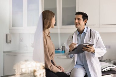 Neşeli erkek doktor teşhisi tartışıyor, hastane ya da klinik ziyareti sırasında genç kadın hastaya sağlık durumu, semptomlar ya da endişeler hakkında sorular soruyor. Sağlık hizmetleri, olumlu haberler, tıbbi danışmanlık